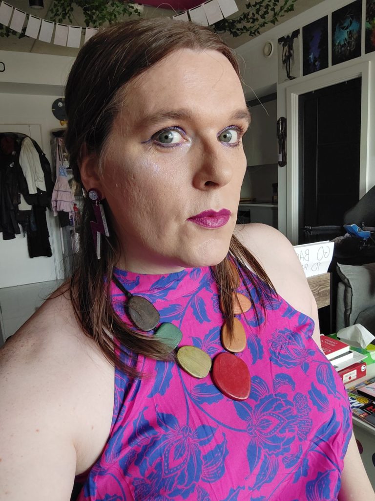 Ashleigh is pictured with shoulder length brown hair, wearing a bright pink and purple top with floral design on it, she is looking at the camera.