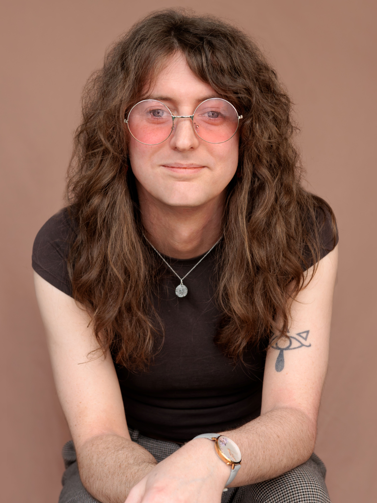 Amber is pictured with thick wavy brown hair that is past shoulder length, and pink sunglasses on, they are looking directly into the camera and are wearing a black tshirt
